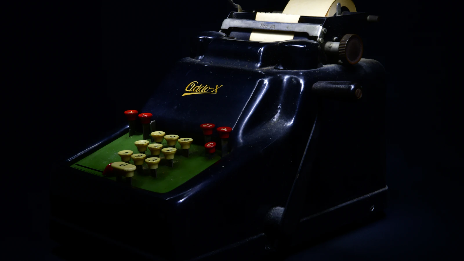 An Addo-X adding machine from around 1950, lit in near darkness with its paper roll on top, totalling only the figures someone keys into it, as an invoice counts only the costs it is shown