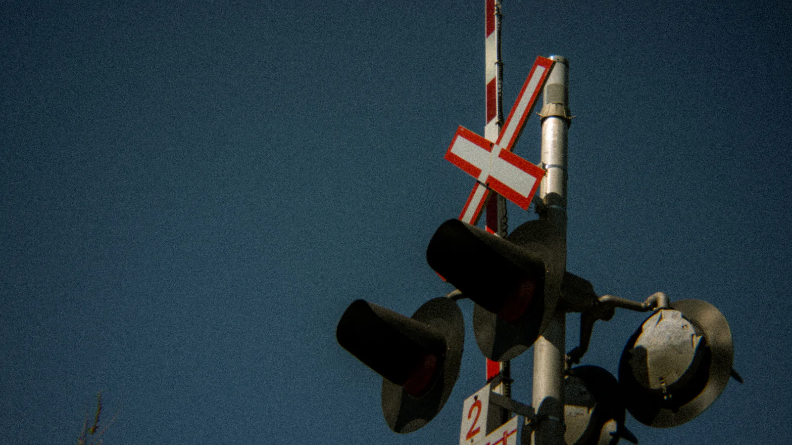 A railway crossing's hooded warning lamps and red-and-white crossbuck against a clear sky, the striped barrier arm standing raised, a signal that warns without closing the road