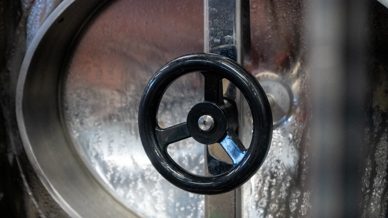 A black handwheel on the sealed round hatch of a stainless steel tank, condensation beaded inside the glass, everything in the vessel kept in until someone turns the wheel