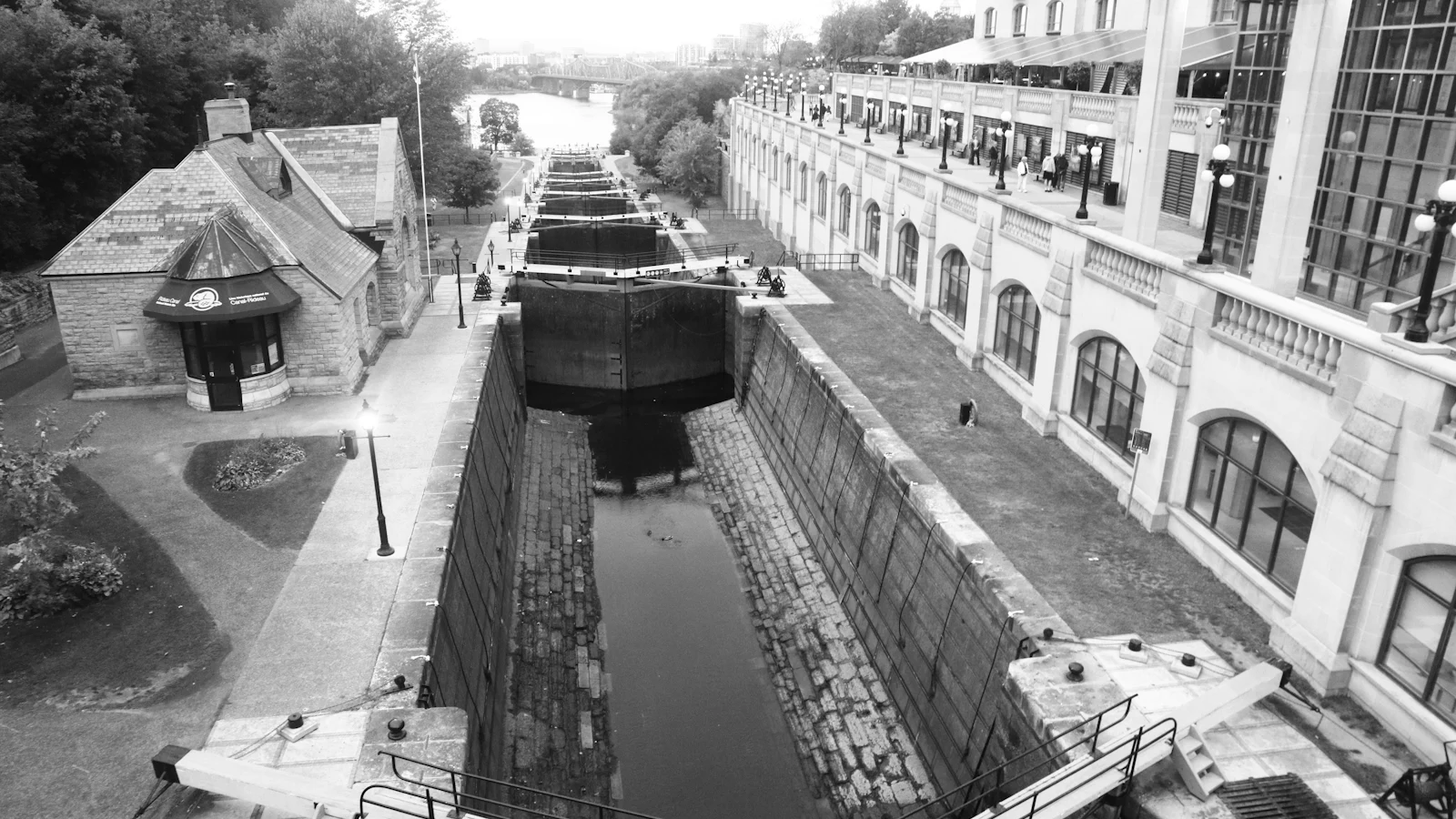 A black-and-white flight of canal locks with closed gates stepping into the distance, each chamber holding a boat until the water level is right before the next gate opens