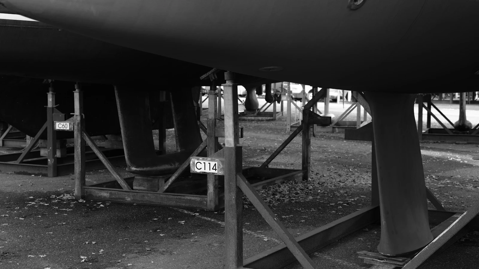 Black and white view beneath boat hulls resting on numbered steel cradles, C60 and C114, ashore in a boatyard: finished, and not yet in the water