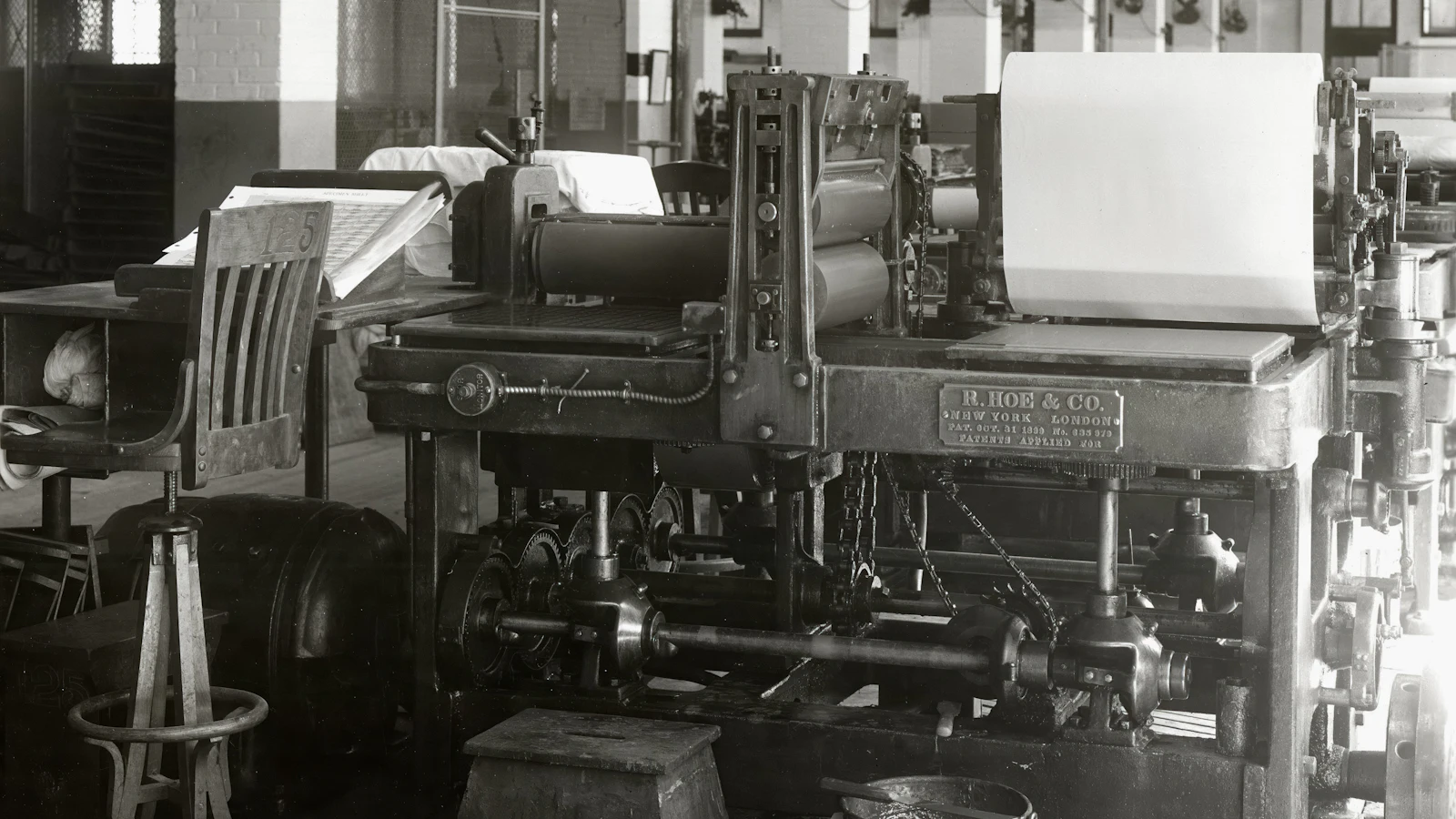 An archival photograph of an R. Hoe & Co. flatbed printing press with an empty operator's chair beside it: the machine runs the job, the people set it up and read the proof