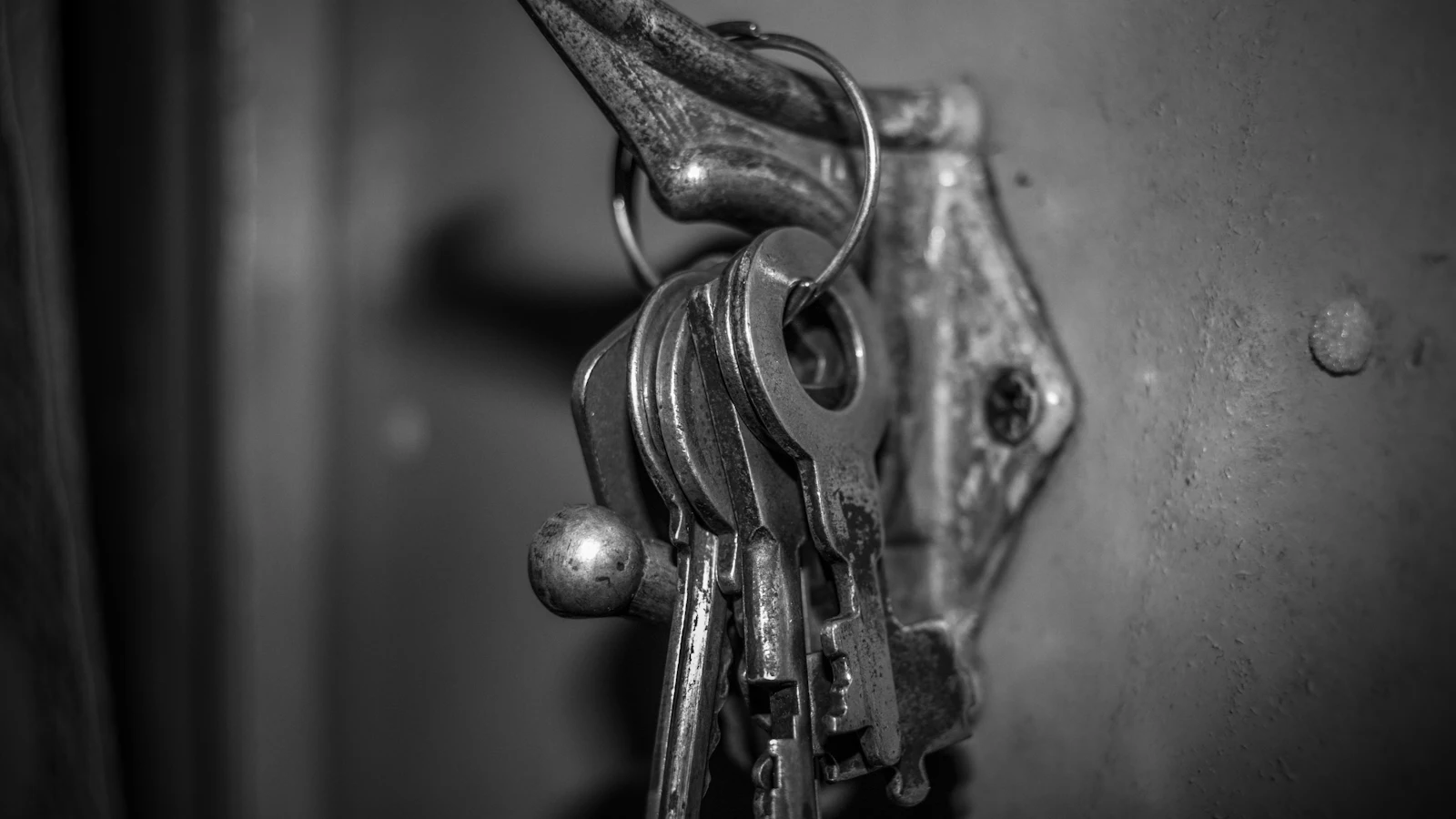 A ring of worn keys left hanging from a metal fitting on a door, in black and white: every key on the ring in reach of whoever passes, the way a long-lived secret sits in an agent's environment