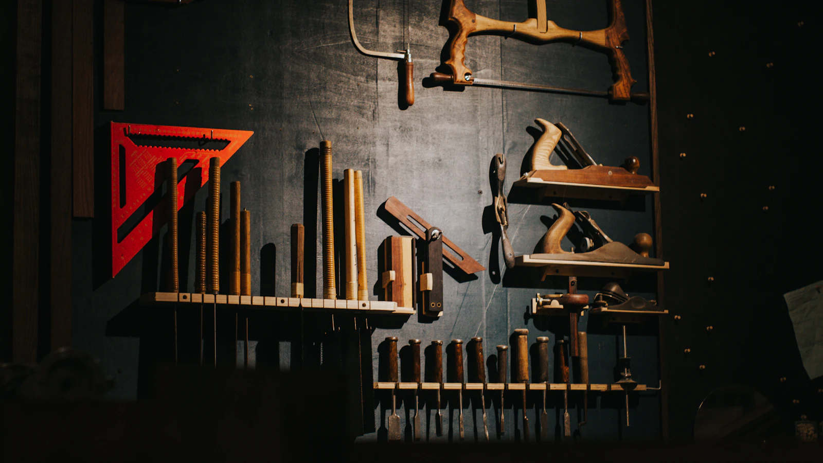 A dark workshop wall where light picks out a bow saw, two planes and a row of chisels while the rest sits in shadow, as agent work puts a few product owner skills in demand