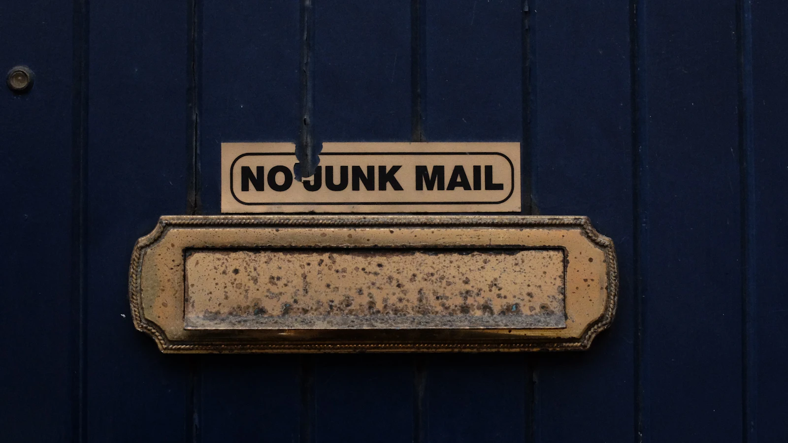 A worn brass letterbox in a dark blue door under a NO JUNK MAIL sign: the sign asks, but the slot still takes whatever anyone pushes through it