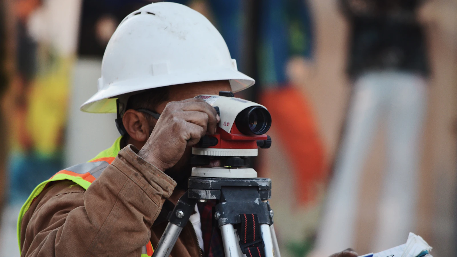 A surveyor sighting through a levelling instrument on its tripod with a notebook in hand, taking the measurement that has to exist before anyone starts building