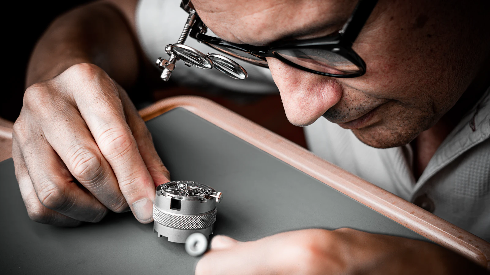 A watchmaker with a flip-down loupe inspecting a movement clamped in a bench holder, the same pair of hands that assembled it now judging it, which is the position a solo founder is in
