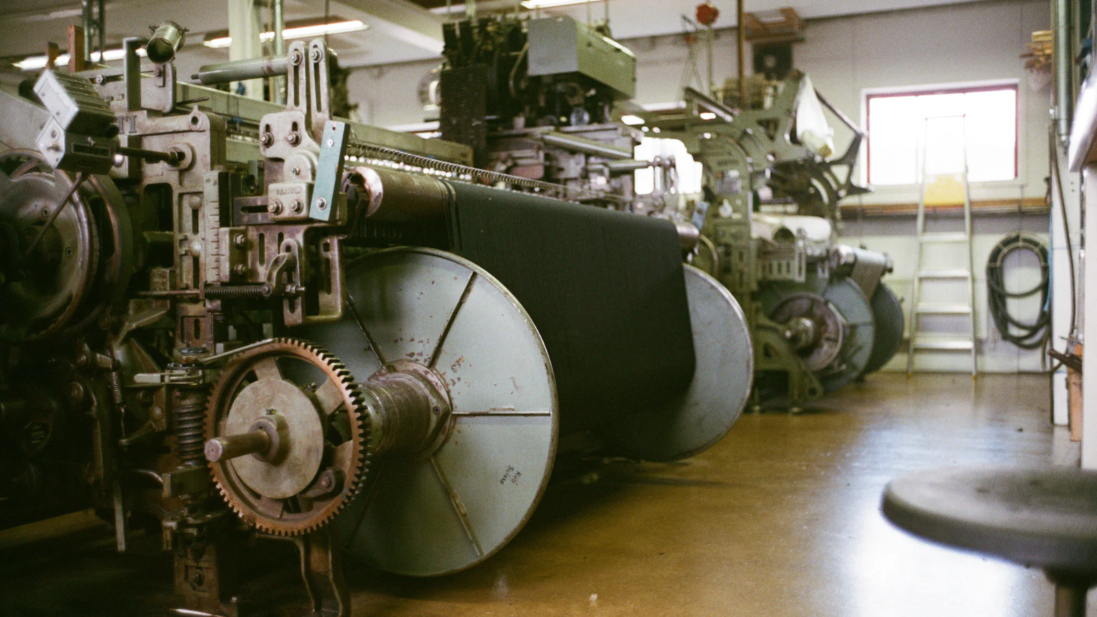 A row of identical industrial looms receding down a workshop floor, cloth rolled on each beam, the first machine's setup repeated down the line