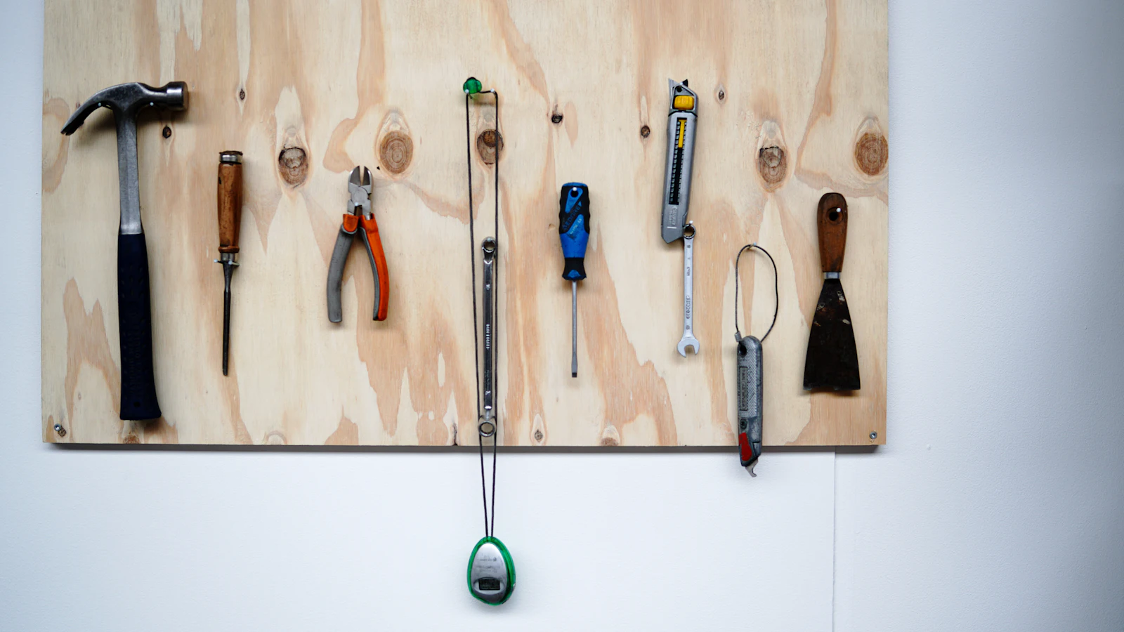 A plywood board on a white wall with everyday hand tools hung in a row, a hammer, pliers, a screwdriver, a knife and a scraper, the tools people already use brought into plain view