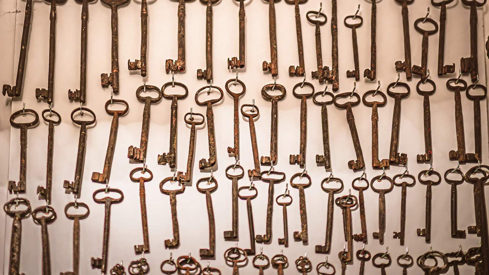 Rows of old iron keys hanging on hooks against a pale wall, none of them labelled, so only the person who hung them knows which door each one opens