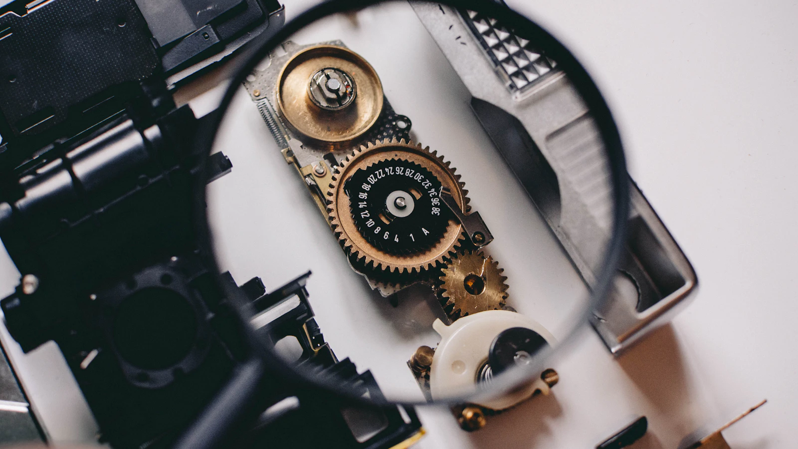 A magnifying glass held over the exposed gear train and frame counter of a disassembled camera, checking the mechanism from outside rather than trusting what it reports