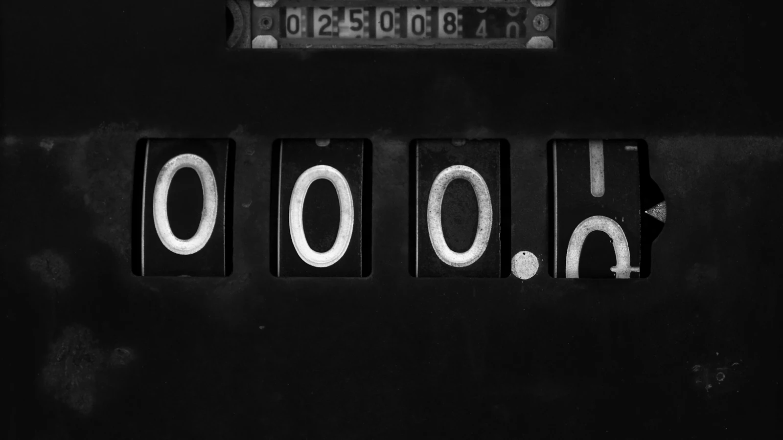 A worn mechanical counter in black and white, its last digit wheel turning over from 0 to 1 below a running total, adding one for each unit finished with no estimate involved