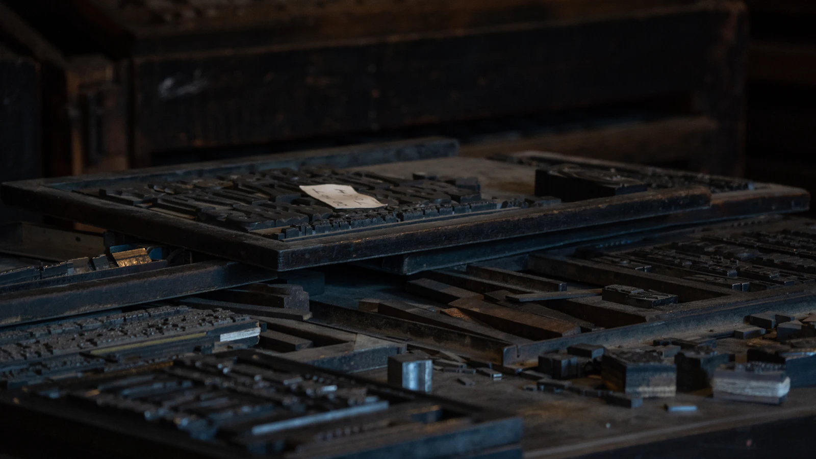 Frames of set metal type stacked in a dark print shop, a paper slip on the top one: text composed letter by letter, ready to print whether or not a word of it is true