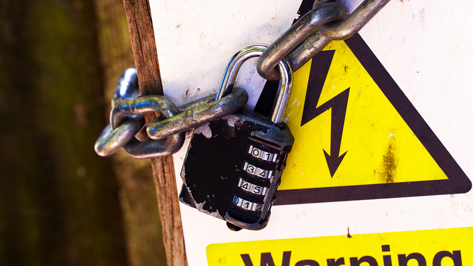 A combination padlock and chain fastened across a yellow high-voltage warning sign, where the sign asks people not to and the chain is what actually stops them